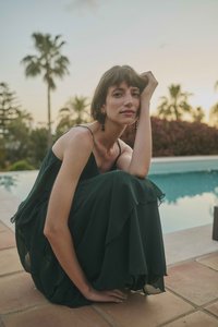 Jeune femme en robe sombre et fluide assise au bord de la piscine au coucher du soleil, reposant sa tête sur sa main, avec des palmiers en arrière-plan.