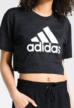 Sort kort Adidas T-shirt med hvidt logo, båret med sorte bukser med snøre, der viser maven og en hånd i lommen.