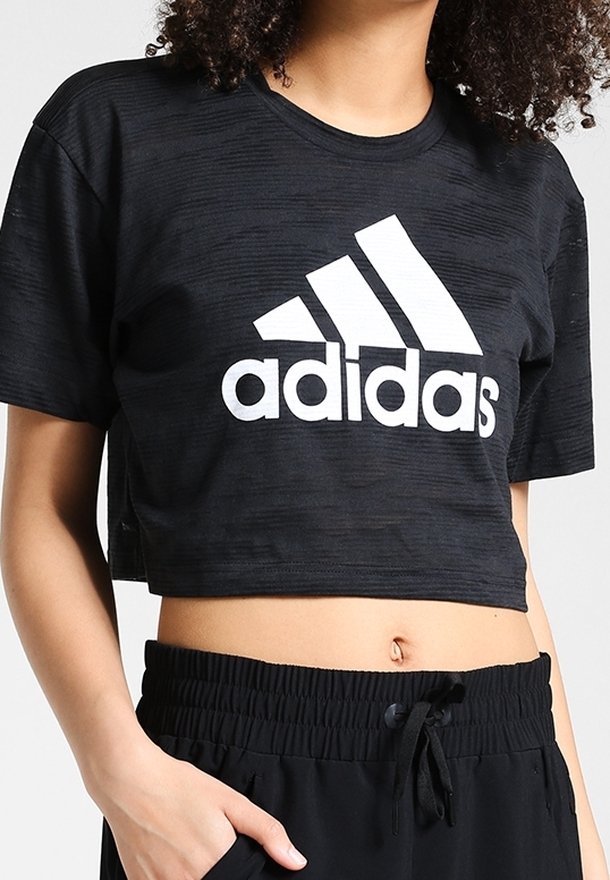 T-shirt noir court Adidas avec logo blanc, porté avec un pantalon à cordon noir, laissant apparaître le ventre et une main dans la poche.