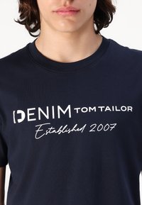 Tumši zila kokvilnas T-krekls ar apaļu kakla izgriezumu, īsām piedurknēm un baltu drukātu tekstu "DENIM TOM TAILOR Established 2007."