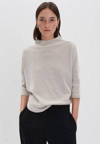 Frau mit glattem braunen Haar und Sommersprossen, die silberne Creolen, ein hellbeiges gestreiftes Oberteil und schwarze Hosen trägt, mit Händen in den Taschen.