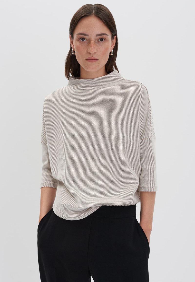 Frau mit glattem braunen Haar und Sommersprossen, die silberne Creolen, ein hellbeiges gestreiftes Oberteil und schwarze Hosen trägt, mit Händen in den Taschen.