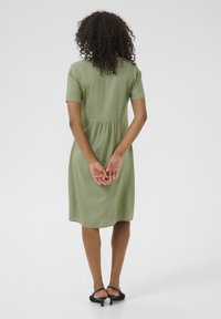 Vrouw met krullend haar staat met haar rug naar voren, draagt een groene jurk tot op de knie met korte mouwen en zwarte lage hakken, met haar handen achter haar samengevouwen.