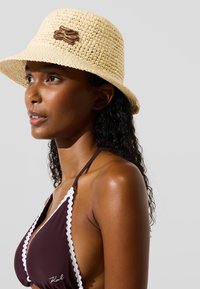 Crème-kleurige geweven strohoed in emmerstijl met een bruin decoratief accent. Model draagt een bruine bikini met witte scalloped rand.