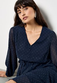 Navy sukienka z długimi rękawami, dekoltem w serek i detalami z guzikami. Posiada białe kropki i ściągniętą talię, co tworzy zwiewny kształt.