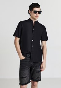 Black short-sleeved button-up shirt with a mandarin collar, paired with distressed dark denim shorts. Model wearing dark sunglasses.