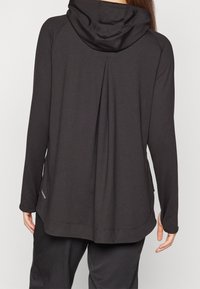 Svart huvlångärmad topp med plisserad rygg, gjord av mjukt material. Har subtila söm- och stickningsdetaljer och en avslappnad passform.