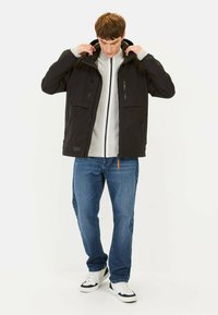 Veste imperméable noire avec capuche, deux poches poitrine avec zippers, une couche intérieure grise, jean bleu et baskets blanches avec accents noirs.