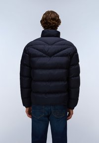 Man met krullend bruin haar, gekleed in een zwarte pufferjas en blauwe spijkerbroek, met zijn rug naar een egale lichte achtergrond.