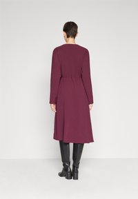 Kvinna iförd en långärmad, knälång klänning i burgundy med ett smalt bälte, tillsammans med svarta knähöga klackstövlar, stående mot en vit vägg.