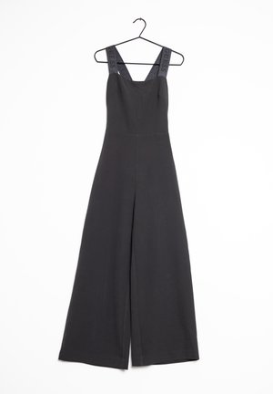 Ärmelloser schwarzer Jumpsuit mit weiten Beinen und breiten Schulterträgern, auf denen das Wort "GUESS," steht, hängend auf einem Kleiderbügel vor weißem Hintergrund.