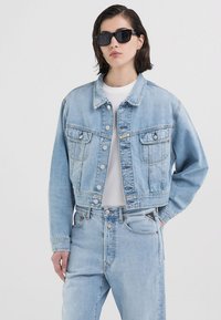 Lyseblå denim cropped jakke med to frontlommer, knappelukking og spiss krage, kombinert med matchende denimbukser og en hvit t-skjorte.