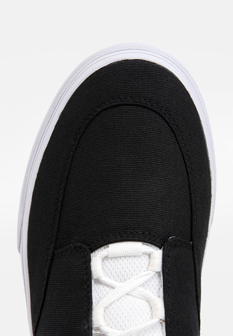 Zapatilla de lona negra con suela de goma blanca, parte superior texturizada y cierre de cordones con lengüeta de malla transpirable.