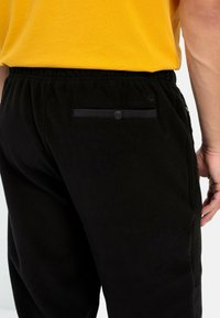 Sorte fleece joggers med elastisk talje og en baglomme med en sort snapknapdetalje, parret med en gul t-shirt.