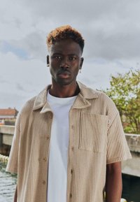 Jeune homme aux cheveux courts et bouclés teints en brun sur le dessus, portant une chemise beige texturée à manches courtes par-dessus un t-shirt blanc, debout en plein air au bord de l'eau.