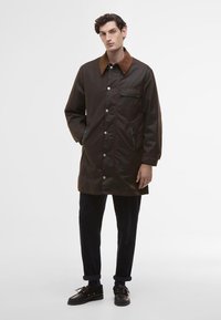 Barna vízálló kabát barna szövetgallérral, nyomógombos záródással, oldalsó zsebekkel és egy mellzsebbel, sötét nadrággal és cipővel párosítva.