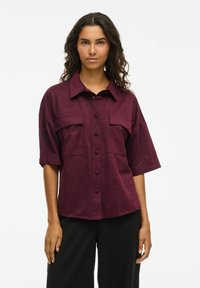 Bordō īsroku krekls no mīksta auduma ar apkakli, priekšējo pogu aizdari un divām krūšu kabatām ar brīvu piegriezumu.