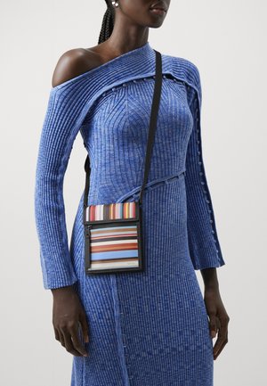 Kobieta w niebieskiej dzianinowej sukience prążkowanej z guzikami i asymetrycznym dekoltem odsłaniającym jedno ramię, niosąca małą wielokolorową torebkę na pasku przez ramię.