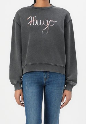 Osoba ubrana w wyblakłą czarną bluzę z różowym, kursywnym napisem „Hugo” oraz niebieskie dżinsy, stojąca na białym tle.