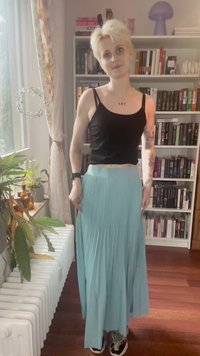 Lühikeste blondide juustega inimene, kellel on seljas must särk, helesinine plisseerimata seelik ja spordijalatsid, seisab ruumis, kus on raamaturiiulid ja taimed.