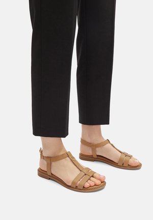 Sandalias de cuero marrón claro con un diseño de tira en T, punta abierta y hebilla en el tobillo. Suelas planas y múltiples correas horizontales que atraviesan el pie.