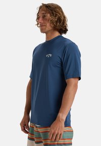Camiseta de rendimiento de manga corta azul, hecha de un tejido suave y ligero. Presenta un pequeño logo blanco en el pecho y un diseño de cuello redondo.