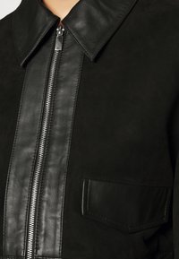 Giacca nera in combinazione di pelle e camoscio, con chiusura a zip, colletto ampio e una tasca a toppa sul davanti.