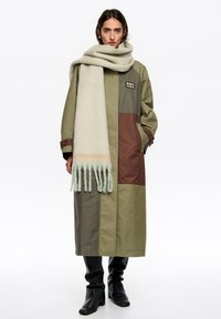 Długi, oliwkowozielony płaszcz z brązowymi i ciemnozielonymi panelami; zestawiony z jasno szalikiem z frędzlami i delikatnymi pastelowymi paskami.
