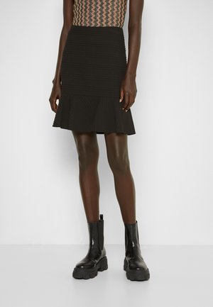 Kobieta w czarnej, teksturowanej mini spódnicy z rozkloszowanym dołem, w połączeniu z czarnymi masywnymi botkami, na jednolitym białym tle.