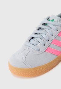 Pelēks zamšādas sporta apavu ar rozā svītrām, fakturētu gumijas zolīti un gaiši zilām šņorēm. Aprīkots ar polsterētu apkakli un klasiskām logotipa detaļām.