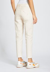 Pantalones de algodón beige con un ajuste estrecho, que presentan bolsillos traseros con una solapa diagonal, combinados con zapatillas blancas y naranjas.