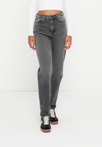 Personne portant un jean gris taille haute, un top blanc à manches longues court et des baskets noires avec des lacets blancs, debout devant un fond blanc.