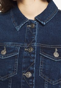 Tummansininen denimmittari, jossa on nappikiinnitys, kaksi rintataskua nappiflapilla ja kontrastia ompelua saumoissa ja kauluksessa.