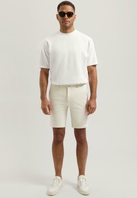 Tricou alb cu mânecă scurtă, asortat cu pantaloni scurți bej deschis, croiți, și adidași albi. Modelul poartă ochelari de soare negri, evidențiind un look casual.