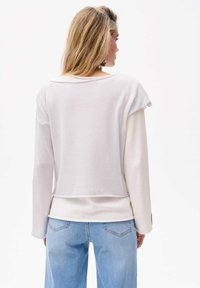 Hellgrauer, ausgestellter Cropped-Sweatshirt mit lockerer Passform, gerippter Textur, weitem Ausschnitt und überschnittenen Schultern, kombiniert mit blauen High-Waist-Jeans.
