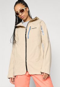 Μπεζ αδιάβροχο μπουφάν με κεντρικό φερμουάρ, κουκούλα και δύο πλαϊνές τσέπες. Διαθέτει μπλε λεπτομέρειες στο φερμουάρ και κεντητό λογότυπο στο στήθος.