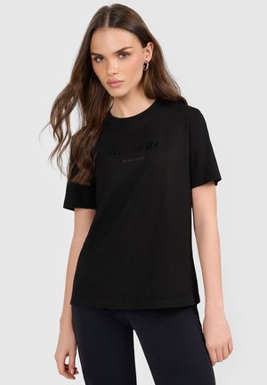Młoda kobieta z długimi brązowymi włosami, ubrana w czarny t-shirt "St. Moritz 4F Ski Club" i czarne spodnie, stoi na tle jednolitego jasnego tła.