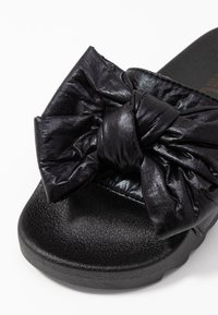 Black slide sandal with a textured sole and wide shiny leather strap tied in a large twisted bow across the top.
