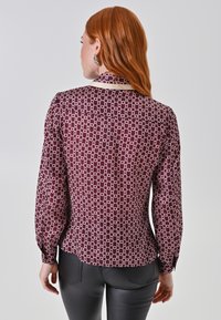 Blusa bordeaux a maniche lunghe con un motivo geometrico bianco; presenta un colletto rotondo, polsini con bottone e un orlo leggermente svasato.