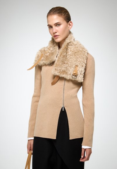Donna che indossa un maglione beige con zip, una sciarpa in shearling color sabbia chiara e pantaloni neri, mentre tiene una tracolla di una borsa color sabbia.
