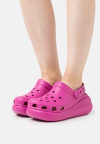 Růžové dřeváky s ventilačními otvory, zvýšenou podrážkou a nastavitelným páskem na patě, vyrobené z lehkého texturovaného materiálu.