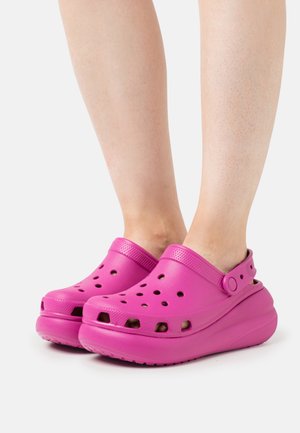 Zuecos rosas con orificios de ventilación, suela elevada y correa trasera ajustable, fabricados en un material ligero y texturizado.