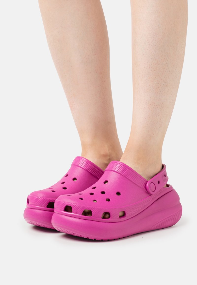 Růžové dřeváky s ventilačními otvory, zvýšenou podrážkou a nastavitelným páskem na patě, vyrobené z lehkého texturovaného materiálu.