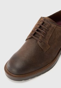 Brown leather shoe with a smooth finish, rounded toe, and textured sole. Features lace-up design and subtle stitching accents.