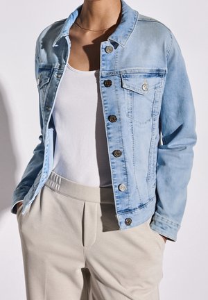 Personne portant une veste en denim bleu clair par-dessus un débardeur blanc avec un pantalon beige, mains dans les poches, sur fond uni.