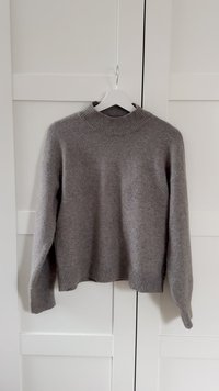 Pilkas megztinis iš mezginio, su ribbingo audinio aukštu kaklu, nuleistais pečiais ir plačiomis rankovėmis. Turi laisvą siluetą ir subtiliai tekstūruotą audinį.