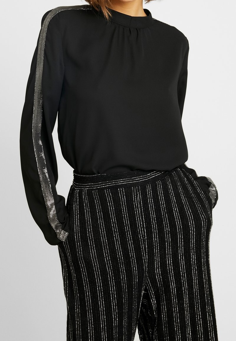 Blouse noire à manches longues avec un col haut, ornée d'accents argentés le long des manches. Associée à un pantalon rayé noir et argent.