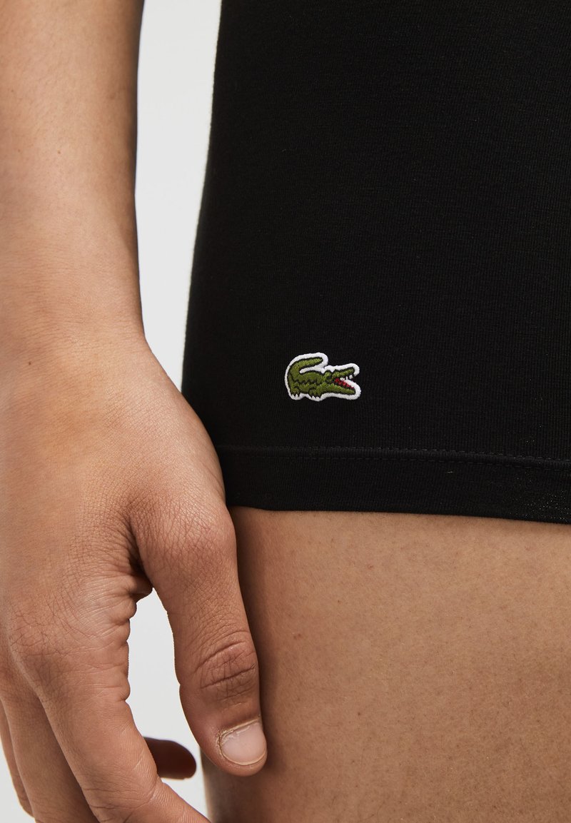 Schwarze Stretch-Shorts mit engem Schnitt, versehen mit einem kleinen gestickten grünen Krokodil-Logo an der Seite und einer glatten Stoffoberfläche.