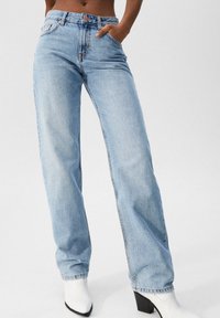 Jeans de tiro alto en azul claro con corte recto, que cuentan con bolsillos frontales y cierre de botón, combinados con botas blancas de tacón.