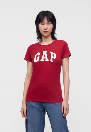 Person står iført en rød T-shirt med korte ærmer med store hvide GAP-bogstaver og blå jeans foran en ensfarvet lys baggrund.